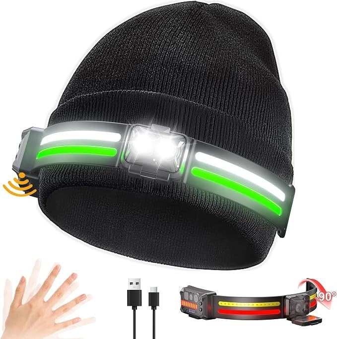 A black beanie features a front-mounted light with a white LED and two green accent lights, designed for hands-free illumination. Visible are also a USB charging cable and an adjustable strap for securing the light.