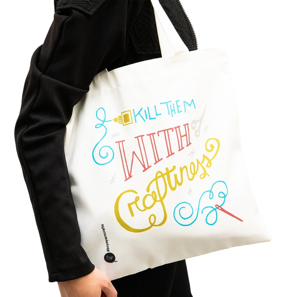 Canvas tote bag featuring a colorful design that reads "Kill Them With Craftiness," held by a person wearing a black jacket. The bag is white with decorative graphics in red, blue, and yellow, perfect for carrying knitting supplies.