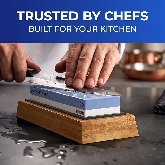 A person's hand holds a knife against a blue Sharp Pebble whetstone labeled with 1000 and 6000 grit, positioned on a bamboo base, with water droplets on the stone and countertop. The background features kitchen utensils and a copper pot.