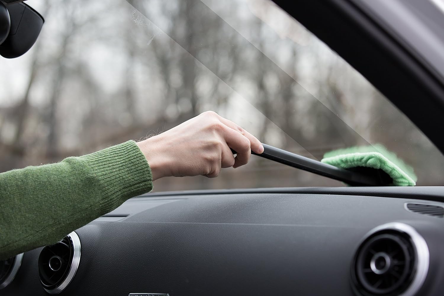 A person uses the EasyWipe Microfiber Window Cleaner with a pivoting head, wiping a car's interior windshield, while wearing a green sweater. The scene shows the cleaner's green microfiber head and black handle, with the car's dashboard visible below.