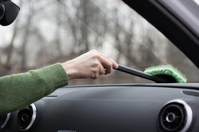 A person uses the EasyWipe Microfiber Window Cleaner with a pivoting head, wiping a car's interior windshield, while wearing a green sweater. The scene shows the cleaner's green microfiber head and black handle, with the car's dashboard visible below.