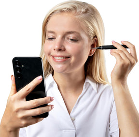 A woman with long blonde hair smiles while holding a black smartphone in one hand and an ear wax removal tool in the other, positioned near her ear. She is dressed in a white shirt, highlighting a clean, modern look.