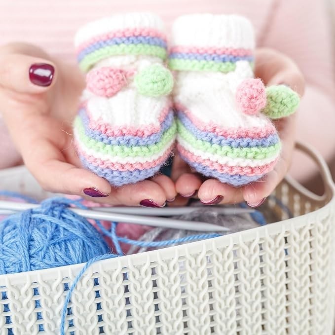 Handcrafted knitted baby booties in soft white, pink, green, and blue colors, featuring pom-pom accents, held in hands above a basket containing blue yarn and knitting needles.