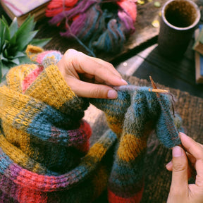 A person is knitting a colorful scarf with various shades of yarn, showcasing a close-up of their hands holding the knitting needles. The setting features a wooden table with additional yarn skeins and a mug in the background.
