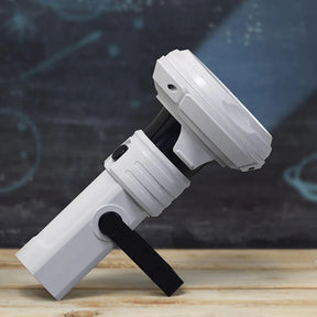 The 3D Galaxy Projector is shown at an angle, featuring a sleek white body with a black section, a handle for easy holding, and a circular projector head. A beam of light illuminates the space, emphasizing its design and functionality.