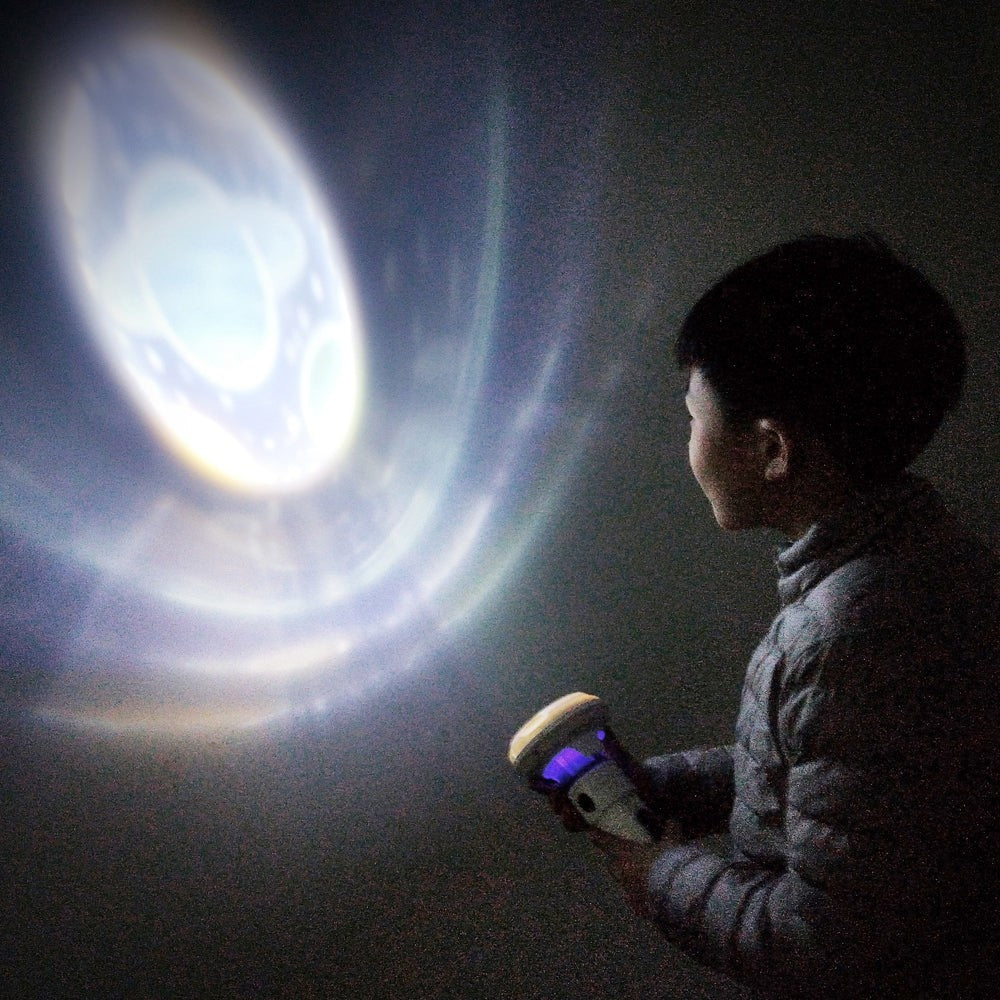 A child wearing a gray jacket is holding the PlaySTEM 3D Galaxy Projector, gazing at a colorful projection of a planet on the wall, with swirling light patterns in the background.