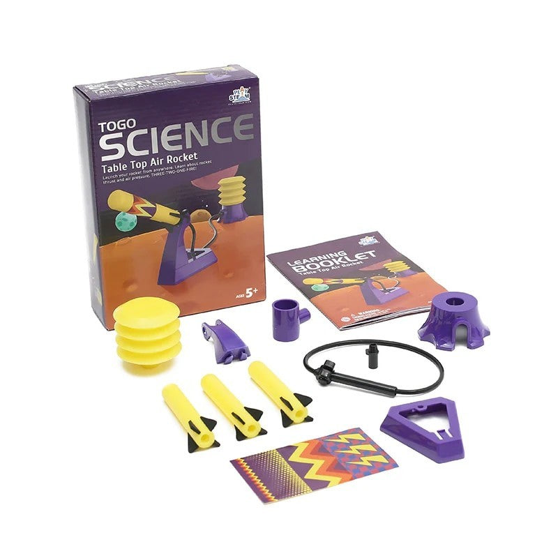 The image features the PlaySTEAM Table Top Air Rocket kit, including a colorful box with illustrations, multiple yellow and purple rocket components, a black launch tube, a learning booklet, and decorative stickers. The components are arranged neatly around the box, showcasing the set's vibrant colors and educational theme.