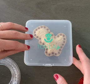 Two sandwiches in fun shapes, one being a bear shape with a decorative border, are placed inside a clear container with a green Tovla Jr. logo. A hand with red nail polish is reaching toward the lid of the container.