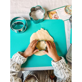 A child’s hands are positioned over a round sheet of cookie dough, using a teal flower-shaped cookie cutter to create a shape. Silver metal cookie cutters of various shapes are visible nearby, along with a teal cutting mat and a recipe card in the background.