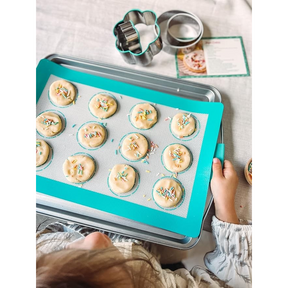 A child’s hands are seen holding a baking tray with unbaked cookie dough discs, each topped with colorful sprinkles, resting on a teal silicone baking mat. Cookie cutters in cloud and round shapes are placed beside the tray, with a recipe card partially visible in the background.