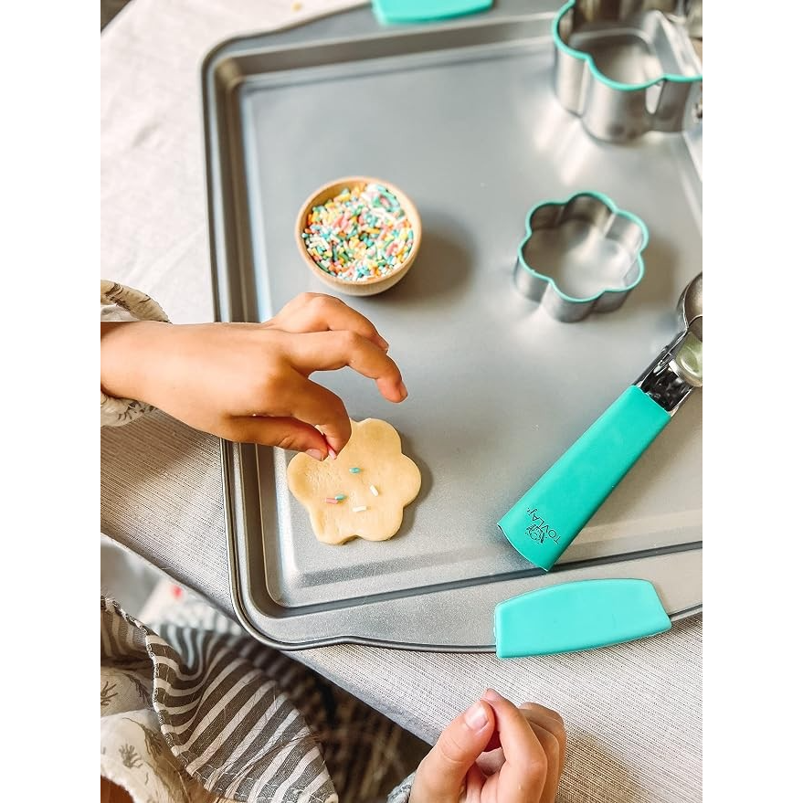 A child's hand decorates a flower-shaped cookie on a baking tray, with a bowl of colorful sprinkles nearby. The tray also features a turquoise ice cream scoop and cookie cutters in various shapes.