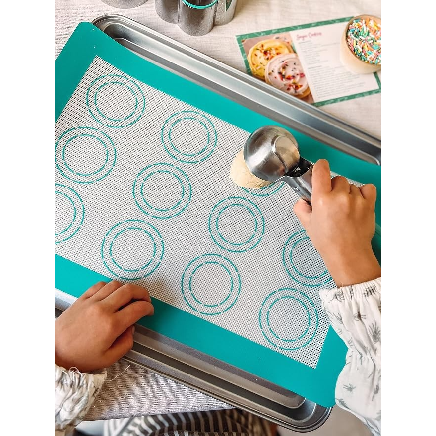 A child’s hand uses a metal scoop to portion cookie dough onto a silicone baking mat with circular outlines on a silver baking sheet, surrounded by additional baking tools and a recipe card.