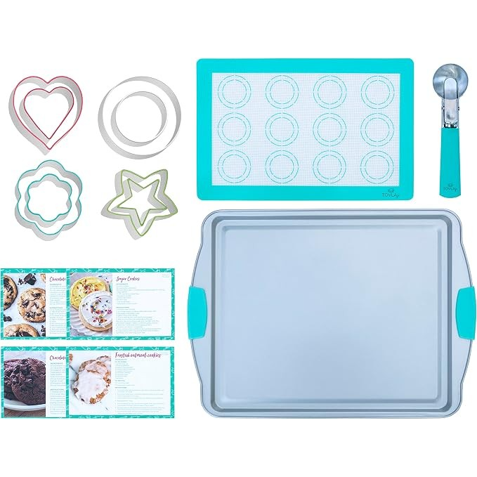 Colorful cookie cutters in various shapes, a blue silicone baking mat with a grid pattern, a metal baking tray, an ice cream scoop, and recipe cards are arranged on a light background, showcasing essential baking tools for kids.