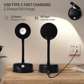 Black wireless motion sensor spotlight on a wooden surface, featuring a round head with a warm light and a base with a touch sensor; one spotlight is positioned near a wall and the other is connected to a power outlet via a USB Type-C charging cable.
