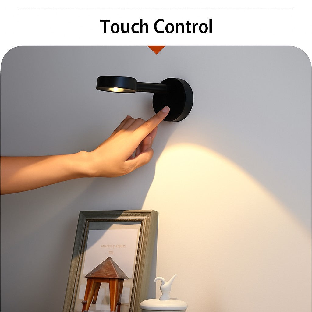 A hand is reaching towards a black wireless motion sensor spotlight mounted on a wall, casting light on a nearby photo frame and decorative items. The setting showcases the touch control feature of the spotlight.