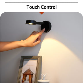 A hand is reaching towards a black wireless motion sensor spotlight mounted on a wall, casting light on a nearby photo frame and decorative items. The setting showcases the touch control feature of the spotlight.