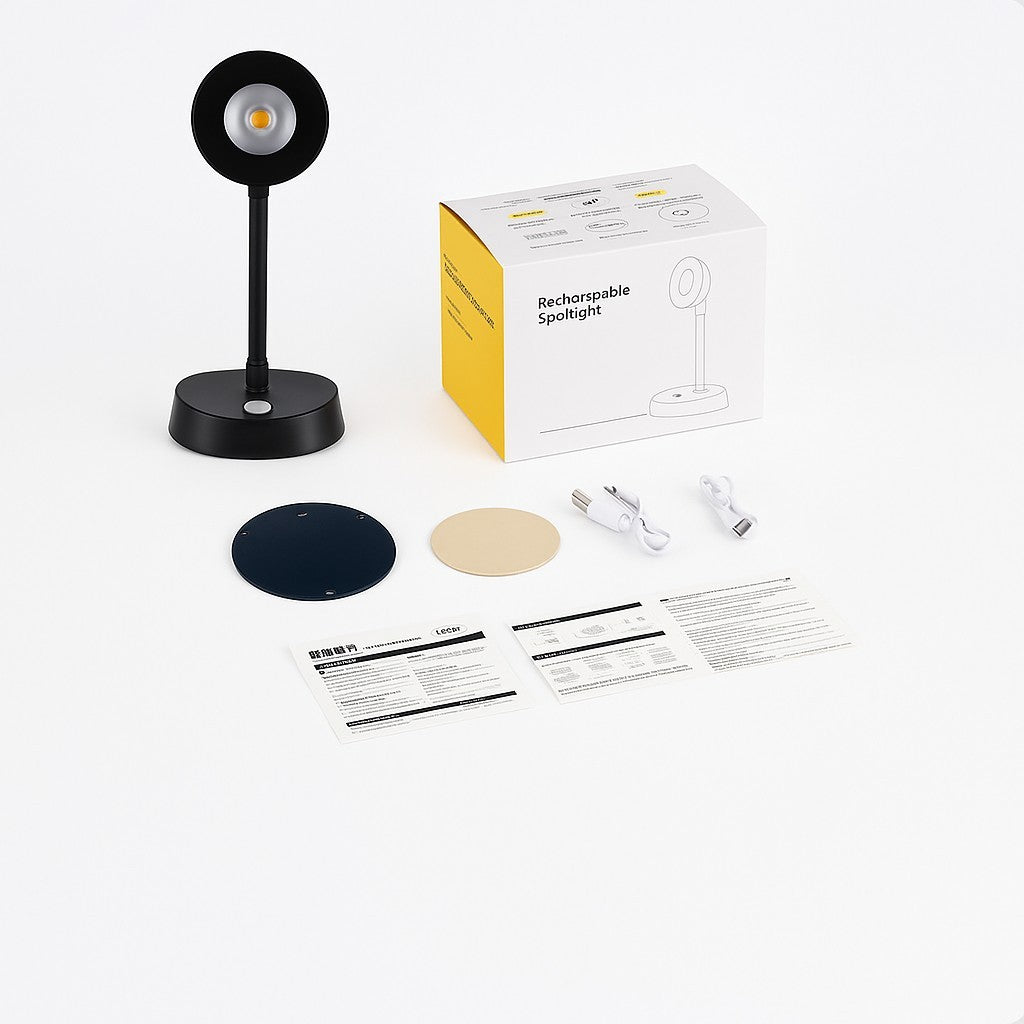 The image shows a black Wireless Motion Sensor Picture Spotlight standing on a round base, accompanied by a yellow and white packaging box, two circular adhesive mounts (one navy blue and one beige), two USB charging cables, and printed instructions.