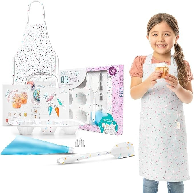 A young girl in a sprinkle-patterned apron holds a decorated cupcake while smiling. The scene includes a baking kit box, various utensils, mixing bowls, and a piping bag, all featuring colorful designs.