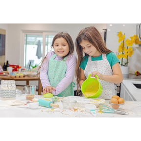 Two children wearing colorful aprons are happily baking in a bright kitchen, with one child rolling dough and the other pouring a mixture from a green bowl. The countertop is adorned with various baking tools, ingredients, and sprinkles.