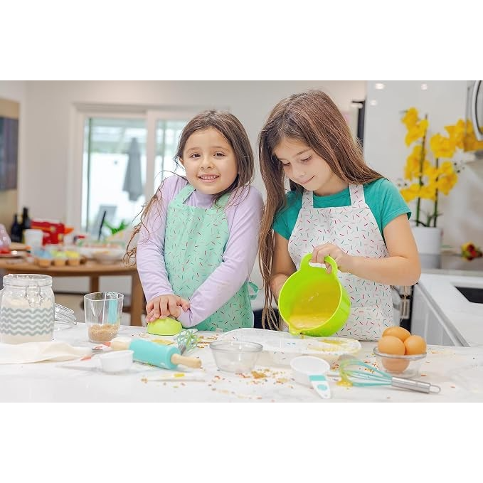 Two children wearing colorful aprons are happily baking in a bright kitchen, with one child rolling dough and the other pouring a mixture from a green bowl. The countertop is adorned with various baking tools, ingredients, and sprinkles.