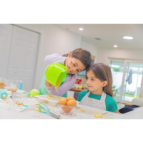 Two young girls are in a bright kitchen, with one pouring batter from a large green mixing bowl into a bowl on the counter while the other watches attentively. The countertop is scattered with baking ingredients, including eggs, utensils, and colorful sprinkles.