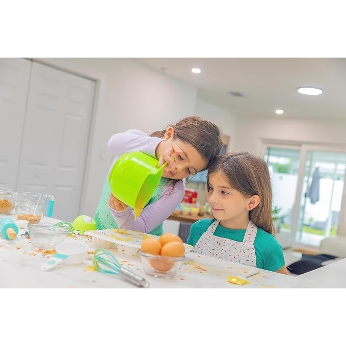Two young girls are in a bright kitchen, with one pouring batter from a large green mixing bowl into a bowl on the counter while the other watches attentively. The countertop is scattered with baking ingredients, including eggs, utensils, and colorful sprinkles.