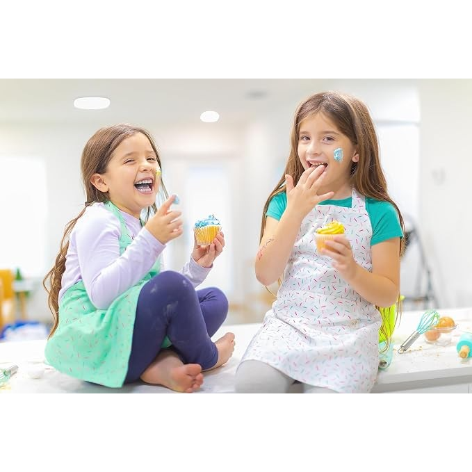 Two girls are seated on a kitchen counter, wearing colorful aprons while laughing and decorating cupcakes. One girl has blue frosting on her face and is holding a cupcake, while the other is playfully eating from her cupcake, surrounded by baking utensils and sprinkles.
