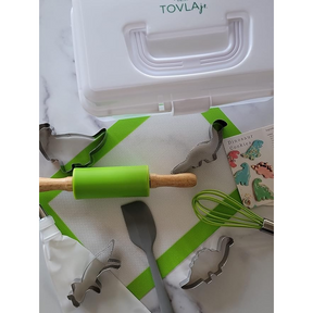A white storage case with "TOVLAjr." on top holds various baking tools, including a green rolling pin, a spatula, a whisk, and dinosaur-shaped cookie cutters, all arranged on a green baking mat next to a recipe card for dinosaur cookies.