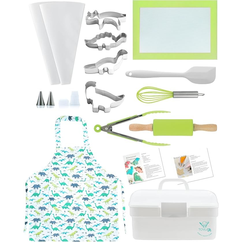 An assortment of baking tools including dinosaur-shaped cookie cutters, a spatula, a whisk, a rolling pin with a green handle, and a white apron featuring a dinosaur print, all arranged neatly with a silicone baking mat and storage container.