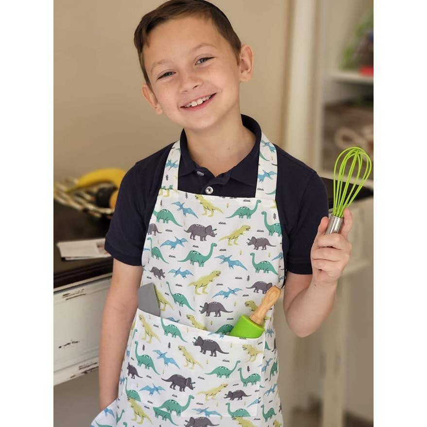 Young boy smiling while wearing a dinosaur-patterned apron, holding a green whisk in one hand and showing baking tools in the apron pocket.