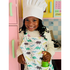 A child wearing a white chef's hat and a colorful dinosaur-patterned apron smiles while holding a green rolling pin, standing in front of a toy kitchen with pink cabinetry.