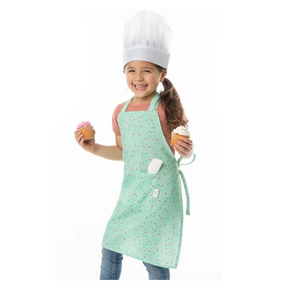 A child wearing a mint green apron with a sprinkle pattern and a white chef hat, holding two cupcakes, one with pink frosting and the other with white frosting. The apron features a front pocket and is tied at the waist.