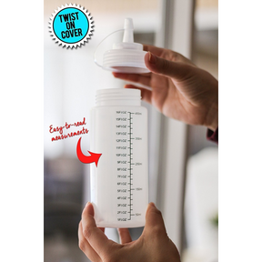 A hand is holding a clear, plastic squeeze bottle with easy-to-read measurement markings on the side, featuring a twist-on cover and an opening for dispensing. The bottle is shown from a slight angle, emphasizing its wide neck and overall design.