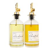 Two glass dispensers filled with light green olive oil and golden peanut oil, each featuring a silver metal pour spout. Each bottle has a label indicating the oil type, enhancing the minimalist design.