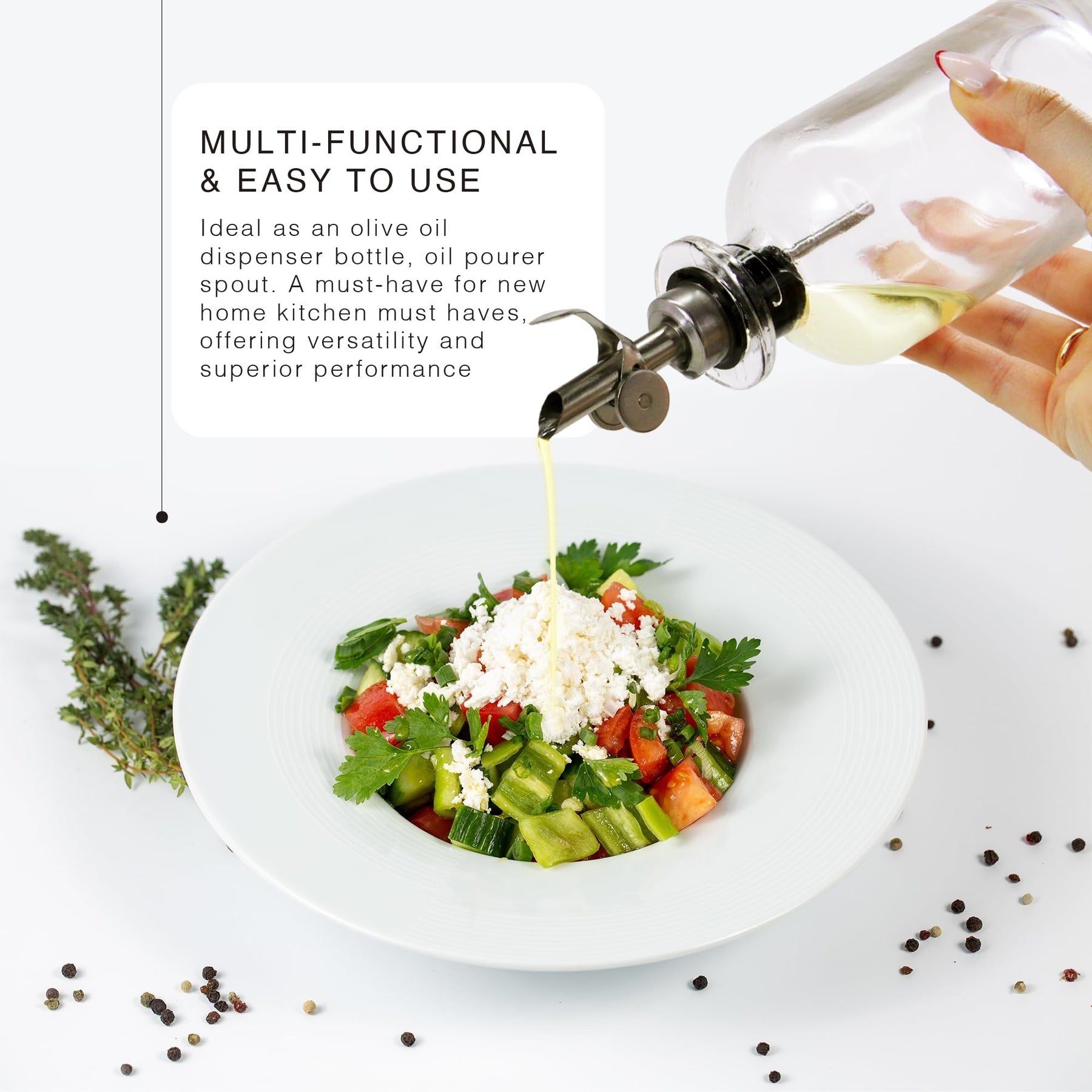 A hand is pouring olive oil from a glass dispenser with a silver metal spout onto a fresh vegetable salad in a white bowl, garnished with feta cheese and herbs, surrounded by various spices on a white surface.