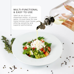 A hand is pouring olive oil from a glass dispenser with a silver metal spout onto a fresh vegetable salad in a white bowl, garnished with feta cheese and herbs, surrounded by various spices on a white surface.