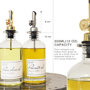 Two glass dispensers filled with olive oil and peanut oil, featuring silver and gold metal pour spouts. Each dispenser has a clear label indicating the contents, with dimensions and capacity information displayed alongside.
