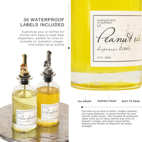 Two glass dispensers, one labeled "Pure Olive Oil" with a gold metal pour spout and the other labeled "Peanut Oil" with a silver pour spout, are displayed on a surface, showcasing their contents. The background includes text about waterproof labels for customization.