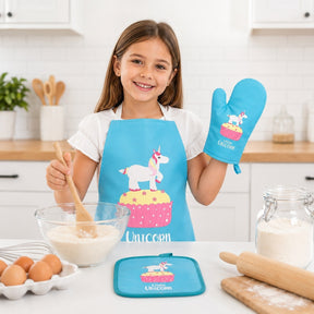 A child wearing a bright blue unicorn apron and matching oven mitt smiles while holding a wooden spoon, mixing ingredients in a glass bowl. The kitchen setting features a countertop with eggs, a rolling pin, and a pot holder with a unicorn design.