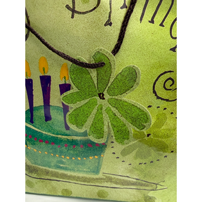 Close-up of a green floral tag attached to the brown handle of a birthday gift bag, featuring a colorful birthday cake design with candles and festive decorations in the background.
