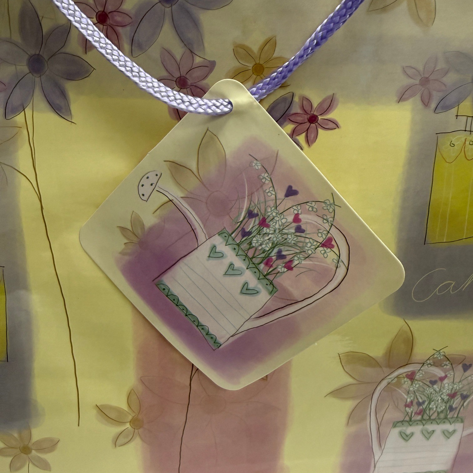 The gift bag features a pastel floral design with purple and yellow hues and a decorative tag showing a watering can and flowers. The bag is finished with lavender handles for easy carrying.