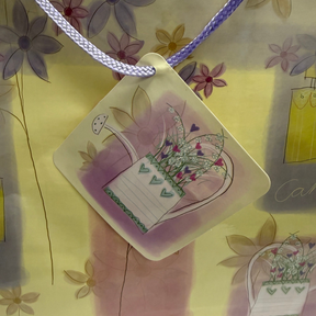 The gift bag features a pastel floral design with purple and yellow hues and a decorative tag showing a watering can and flowers. The bag is finished with lavender handles for easy carrying.