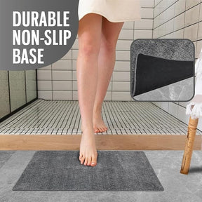 A person is standing on the gray doormat in a bathroom setting, with water droplets on their legs, highlighting its absorbent nature. The inset shows a close-up of the doormat's textured surface and durable non-slip base.