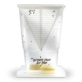 Transparent fruit fly trap with a triangular shape, featuring a slotted top for entry, and containing visible fruit pieces inside. The text "THE ULTIMATE FRUIT FLY TRAP" is printed on the lower part of the trap.