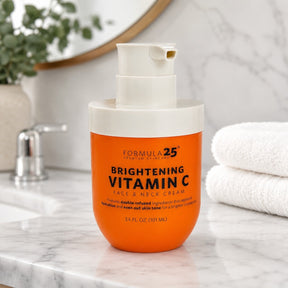Brightening Vitamin C face and neck cream in a vibrant orange jar with a white pump top, displayed on a marble countertop beside neatly folded white towels.