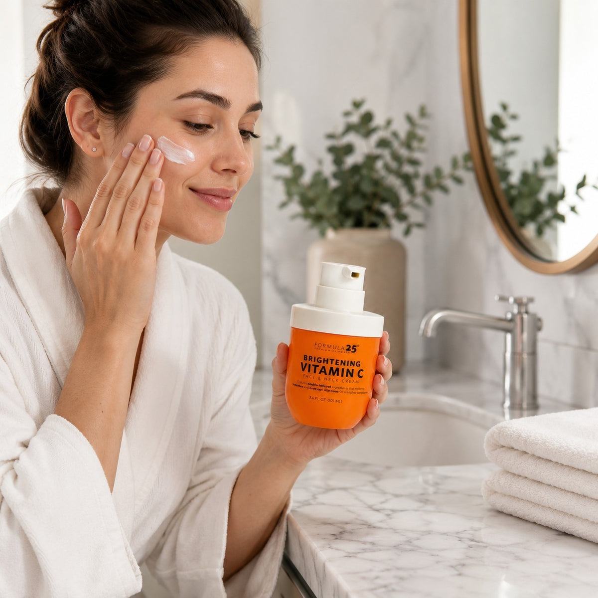 A woman in a white robe applies cream to her face while holding a bright orange jar of Vitamin C Face & Neck Cream at a bathroom sink, with a soft, neutral backdrop and neatly stacked towels nearby.