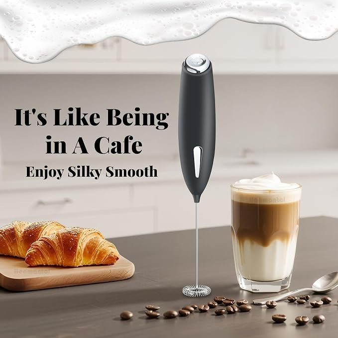 The electric milk blender features a sleek black handle and a stainless steel whisk, positioned next to a frothy coffee drink in a glass, accompanied by croissants and coffee beans on a kitchen counter.