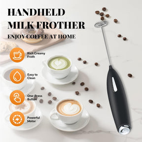 The handheld milk frother features a sleek black design with a slender body and a silver button, positioned next to three cups of coffee, each topped with decorative froth, on a marble surface adorned with coffee beans.