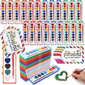 A colorful assortment of heart-shaped watercolor paint sets in various colors, neatly arranged with some sets stacked and a paintbrush shown in use creating a green heart. Includes playful branding cards that read "We've painted a great year together!"