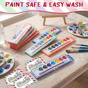 Heart-shaped watercolor paint sets in vibrant colors are arranged on a wooden table, featuring eight hues in each container. Additional paint palettes and brushes are visible, along with cheerful promotional cards highlighting the product.